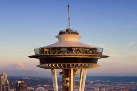 The image shows the top of the Space Needle in Seattle, with a panoramic observation deck crowded with people. In the background, Mount Rainier is visible under a clear blue sky during sunset.