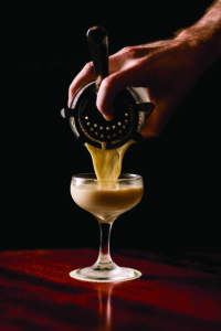 A person's hand pouring a yellow cocktail through a strainer into a stemmed glass on a dark wooden surface, set against a black background.