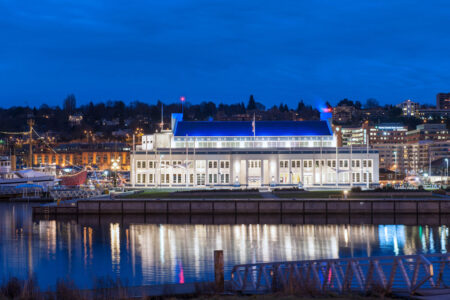 Against a backdrop of city lights at dusk, a waterfront building with bright white lights offers a captivating view—a must-see among things to do in Seattle. A boat is moored nearby, while the deep blue sky and water reflect the luminous scene perfectly.