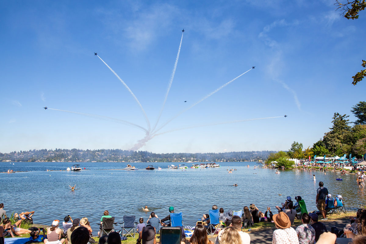 Air show with six jets flying in formation over a lake, leaving smoke trails. Spectators on the grassy shore and people in boats watch the display under clear blue skies.