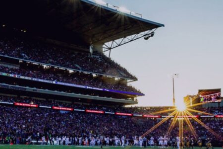 A vibrant football stadium packed with cheering fans under the evening sky. The sun sets, casting a warm glow, while players are on the field under bright stadium lights.