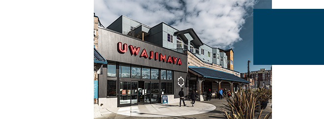 Exterior of Uwajimaya store with pedestrians walking nearby. The building features large glass windows and bold red signage. Modern apartments rise above, set against a partly cloudy sky.