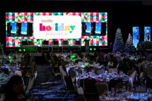 A festive event setup with tables and chairs in front of a vibrant "Seattle Holiday Brunch" stage display. Decorated Christmas trees are on the right, and the atmosphere is lively with dim lighting.