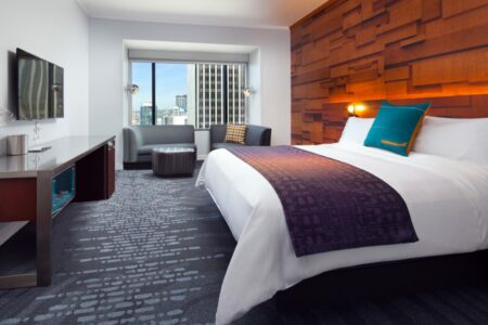 Modern hotel room with a large bed featuring white linens and a purple throw. The room has a textured wood accent wall, a flat-screen TV, and a seating area by a window with a city view. The decor includes colorful pillows.