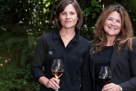 Two women standing outdoors holding wine glasses, surrounded by greenery. Both are smiling and wearing black clothing. The background features a mix of trees and foliage.