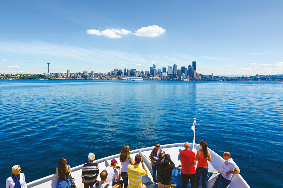 A white Argosy Cruises tour boat filled with people wearing colorful shirts fills the foreground. The blue waters of Puget Sound separate the boat from the city skyline against a blue sky.