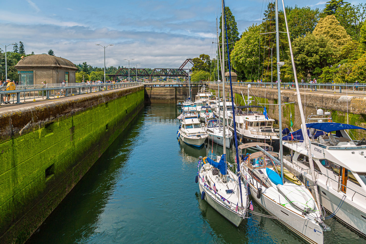 Several boats are docked in a narrow canal with a green, mossy wall on the left and a bridge in the background. Trees and a small building are visible under a blue sky with clouds. Pedestrians walk along paths on either side of the canal.