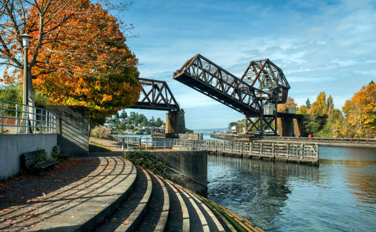A canal scene with an open drawbridge over the water, surrounded by autumn trees with orange leaves, is reminiscent of picturesque sights and things to do in Seattle. Curved stone steps lead down to the canal, and the sky is clear and blue.