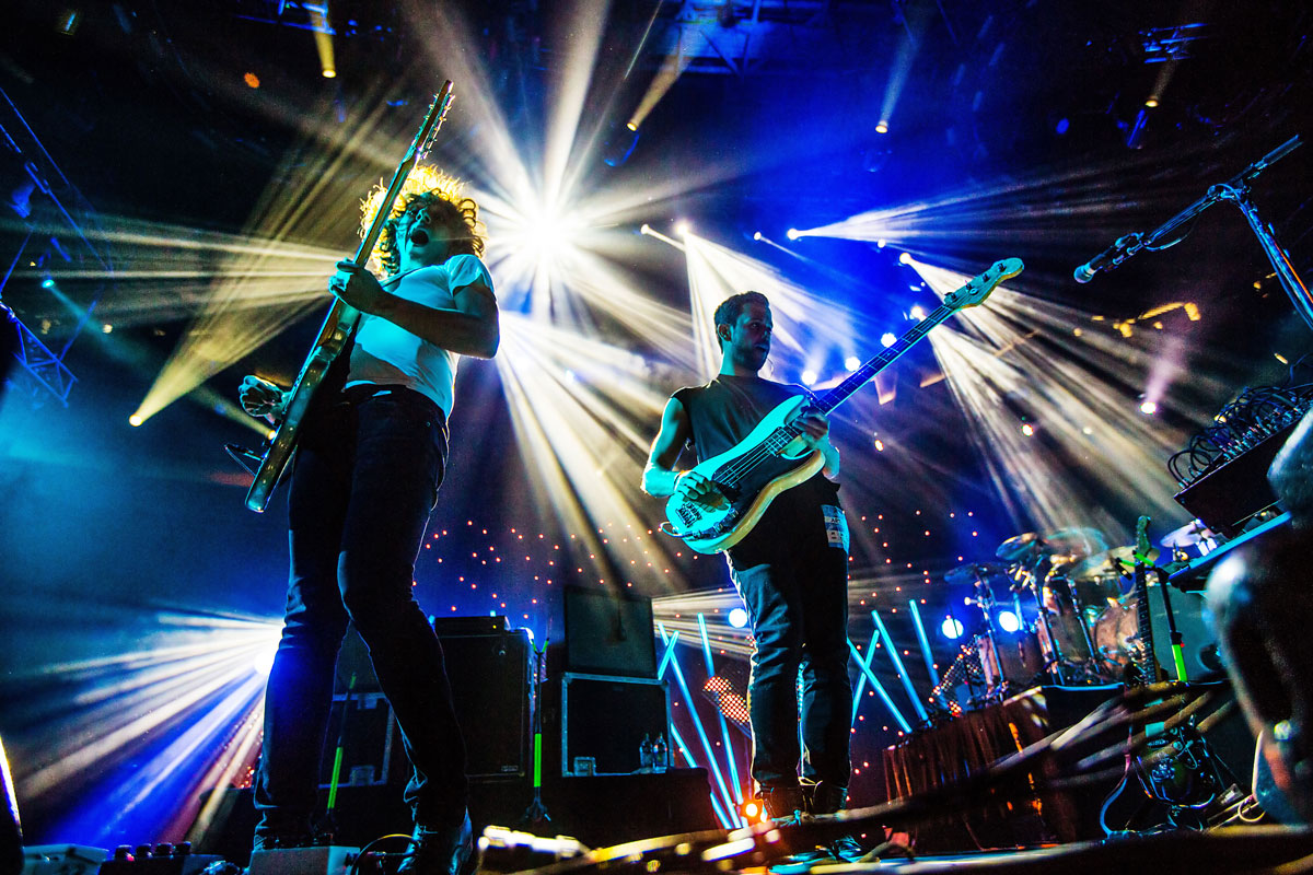 A vibrant concert scene with two musicians playing electric guitars under colorful, bright stage lights. The background is filled with star-like effects, amplifying the energetic atmosphere of the performance.