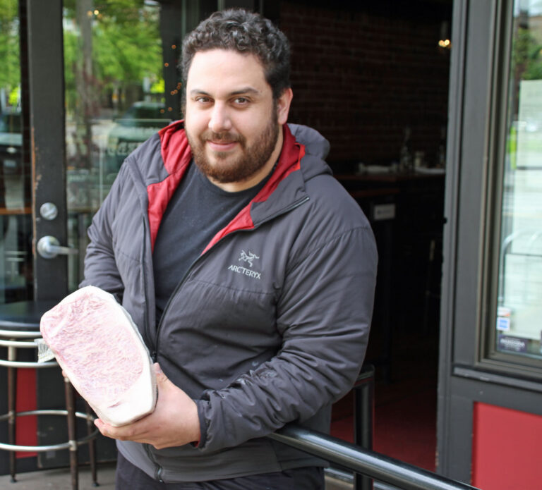 A person with curly hair, wearing a gray jacket with a red lining, is standing outside a building, holding a large piece of marbled meat and smiling at the camera.