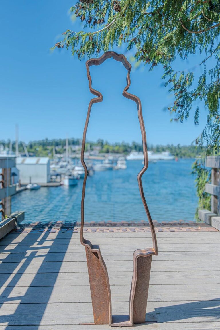 A metal silhouette sculpture of a person stands on a wooden deck overlooking Bremerton's scenic marina, with sailboats and clear blue water in the background. A tree branch extends slightly into the scene from the right, capturing Bainbridge Island’s enchanting maritime essence.