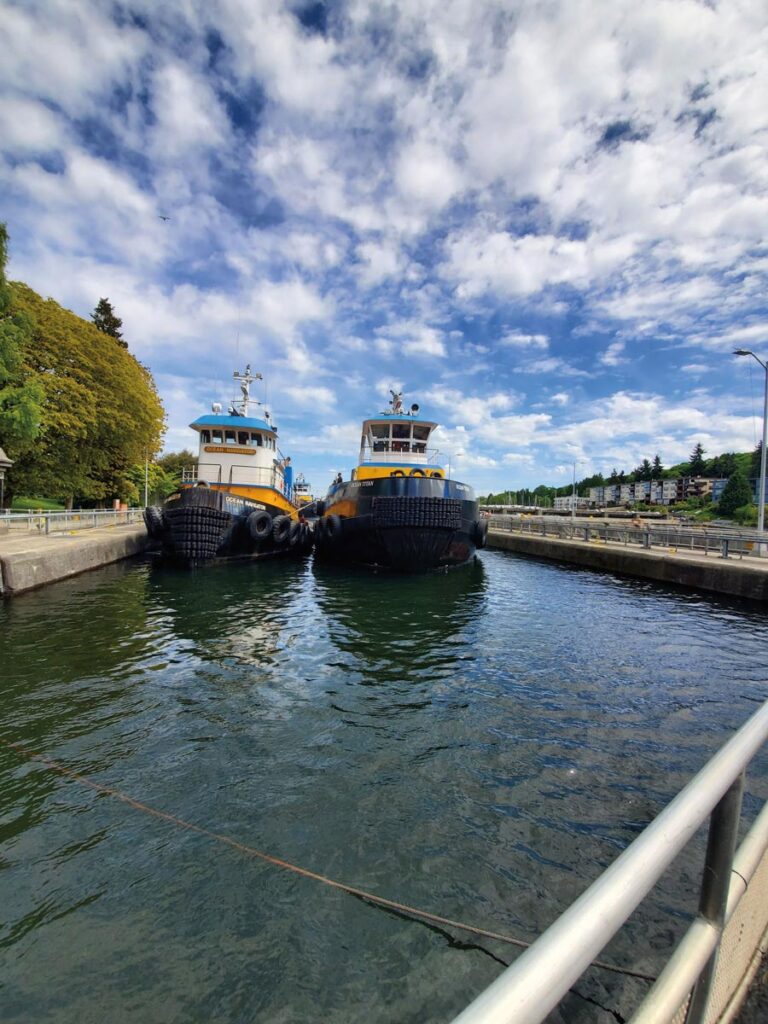 Two tugboats are docked side by side in a canal under a partly cloudy sky. In the picturesque Ballard neighborhood, trees line the waterway, accompanied by a railing and distant buildings, creating a scenic view.