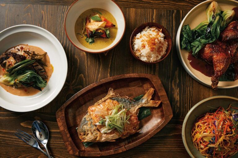 A wooden table with a spread of dishes, including a whole grilled fish on a wooden plate, a bowl of rice, roasted chicken with greens, a dish of mixed vegetables, a curry bowl, and colorful noodle salad. A spoon and fork are placed on the table.