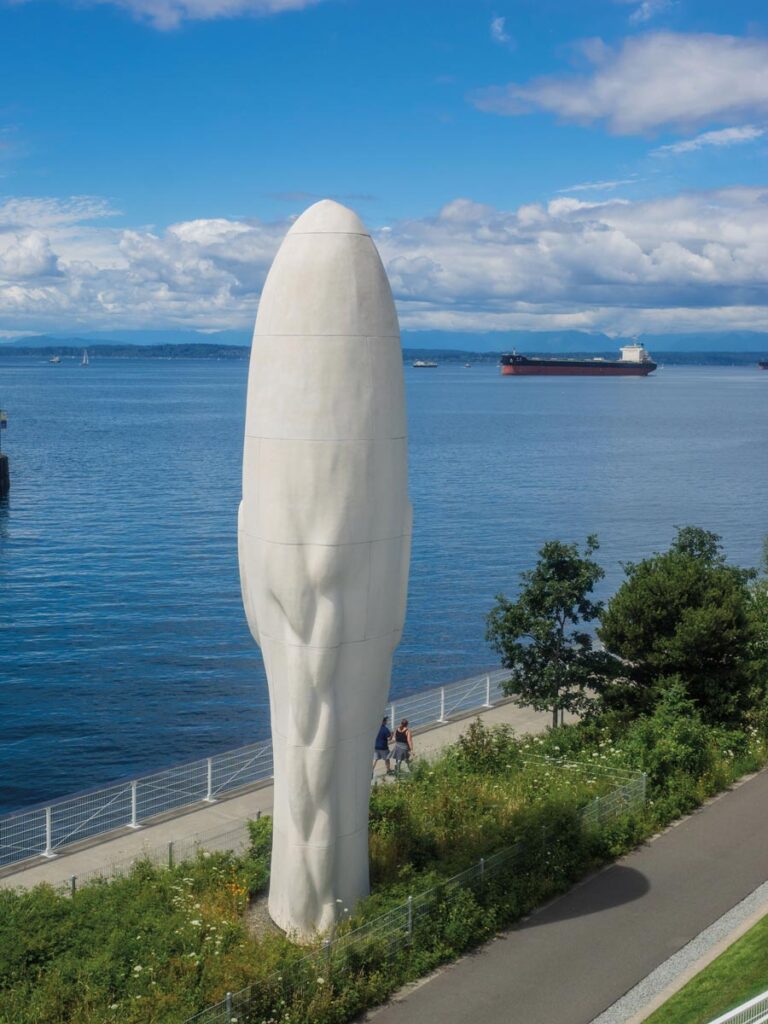 A towering white sculpture of a head graces the waterfront path in the Belltown neighborhood, Seattle, surrounded by lush greenery. The scene is completed with views of a large body of water, a ship in the distance, and a partly cloudy sky.