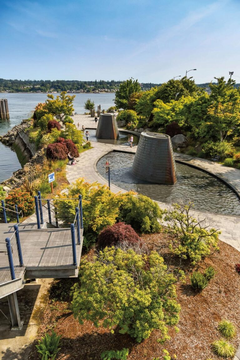 Scenic riverside view of a park with pathways and two large conical water features, reminiscent of Bainbridge Island. People stroll along the paths, surrounded by trees and shrubs, with a body of water and Bremerton's forested shoreline in the backdrop under a clear blue sky.