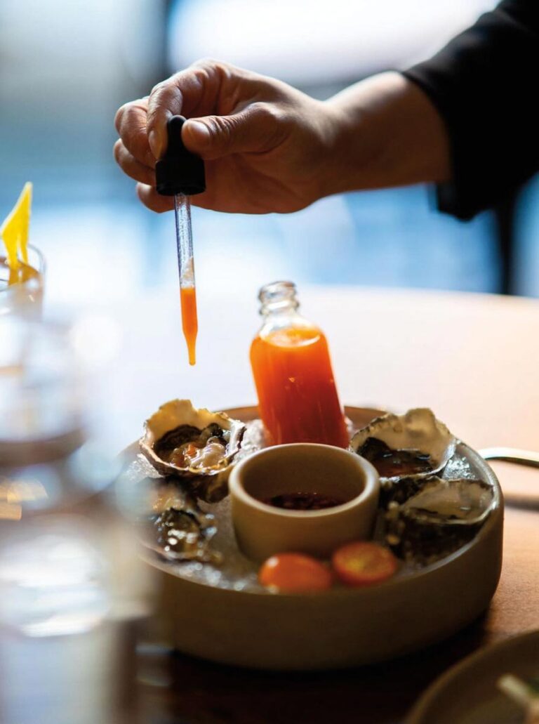 In the cozy ambiance of a Capitol Hill neighborhood eatery, a hand uses a dropper to add sauce to oysters on a plate. Nearby, a small bottle holds extra sauce, and a dish of sliced cherry tomatoes sits ready. The scene is warmly lit atop a rustic wooden table.