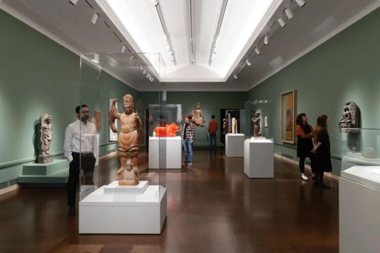 A museum exhibition room in the Capitol Hill neighborhood features sculptures and artifacts displayed in glass cases. Visitors observe and discuss the artwork as dim lighting and green walls create a serene atmosphere.