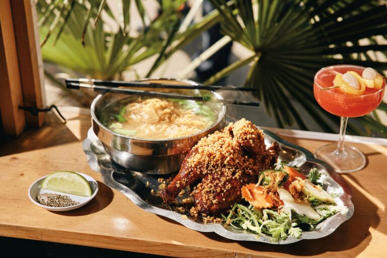 A plate of spicy fried chicken garnished with herbs and vegetables sits next to a bowl of ramen, complete with chopsticks on top, reminiscent of the vibrant flavors found in Chinatown-International District. A cocktail with lemon slices is in the background, set against a backdrop of green leafy decor.