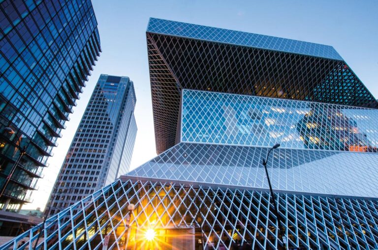 Modern glass buildings in Seattle's downtown neighborhood create a striking cityscape, featuring the distinctive geometric design of the Seattle Central Library with its reflective, grid-like facade. The sun peeks through the lower section, casting a warm glow as other skyscrapers rise in the background.
