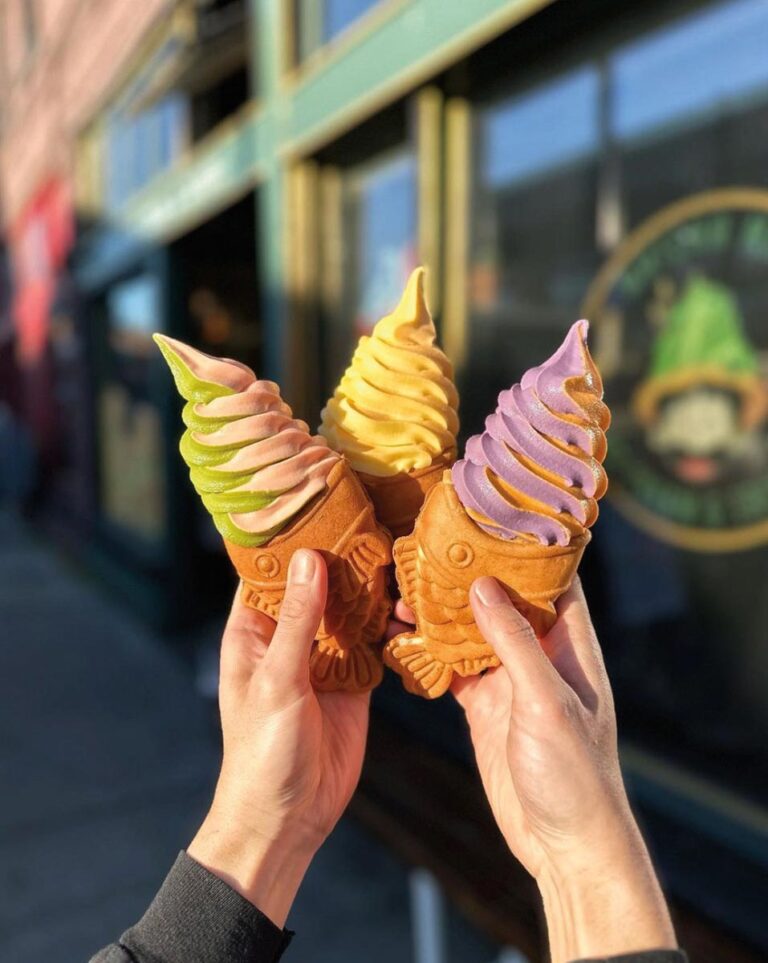 Two hands hold three colorful soft-serve ice creams in taiyaki fish-shaped cones, set against the charming backdrop of a building with a round sign in the vibrant SoDo neighborhood. The creamy mix features pastel green, pink, yellow, and purple hues.