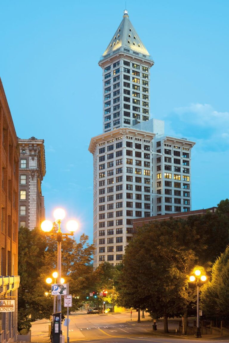 A tall, historic skyscraper with a pointed roof stands illuminated against a clear evening sky in the Pioneer Square neighborhood. It is surrounded by trees and flanked by lit streetlamps and buildings on a quiet street.