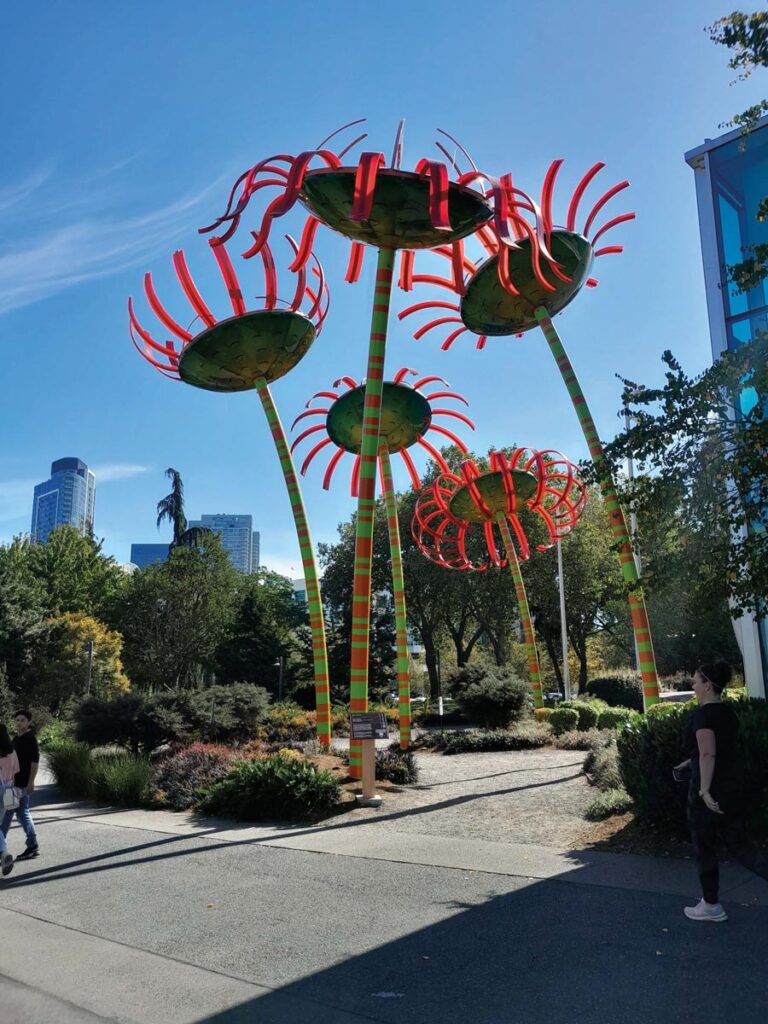 In the Queen Anne neighborhood, colorful, tall sculptures resembling flowers with red petals and green stems grace a park setting, framed by trees and buildings in the background. People stroll along the path under a clear blue sky.