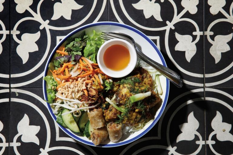 A colorful dish featuring grilled fish covered in herbs, spring rolls, shredded carrots, peanuts, greens, and cucumbers sits stylishly on a black and white patterned surface. This vibrant taste of the South Lake Union neighborhood includes a small bowl of dipping sauce and a fork placed elegantly on the plate.