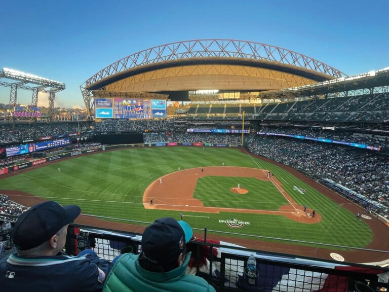 A panoramic view of a baseball stadium, nestled between the vibrant SoDo and Georgetown neighborhoods in Seattle, captures the excitement of a game. The retractable roof is partially open, and the field is brightly lit. Cheering fans fill the stands as large screens display game details.