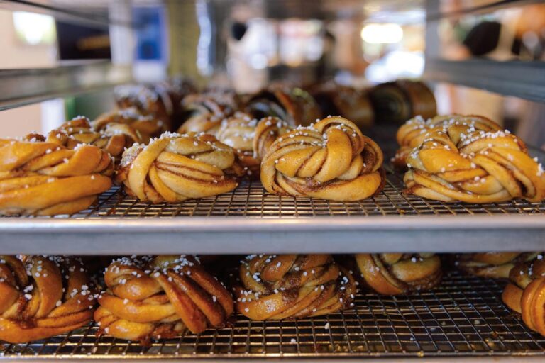 Rows of freshly baked cinnamon buns on metal racks, topped with pearl sugar, evoke the cozy charm of a University District Seattle neighborhood bakery. The buns, with their twisted spiral shape and golden-brown crust, sit against a slightly blurred background that suggests warmth and invitation.
