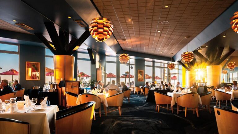 A modern restaurant in Seattle's waterfront neighborhood features large windows showcasing an ocean view. The warm sunset light filters in, illuminating tables set for dining. Orange pendant lights hang from the ceiling, complementing the cozy ambiance of wooden chairs and white tablecloths.