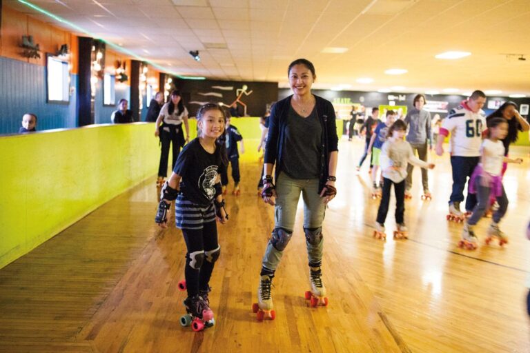 A group of people roller skating indoors on a wooden rink. A woman and a child smiling and skating towards the camera in the foreground. People of different ages skate around them, and the background is lively and well-lit.