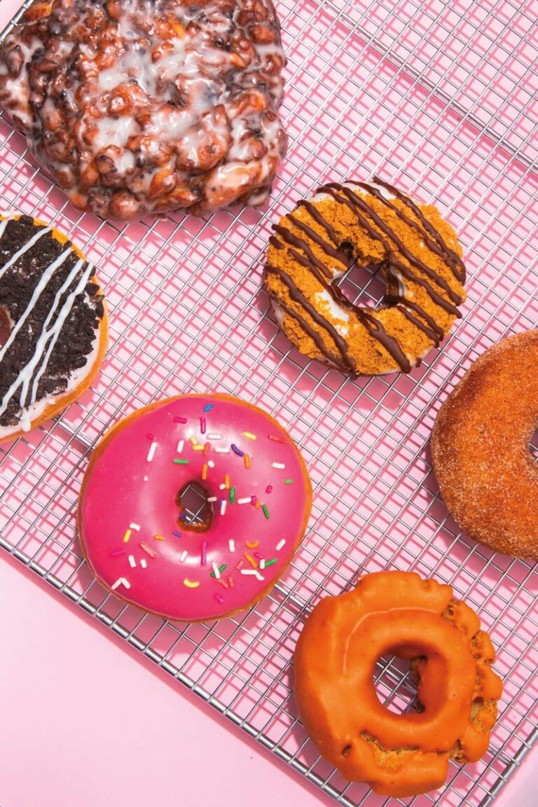 A variety of colorful donuts from the vibrant West Seattle neighborhood sit on a cooling rack. The selection includes a pink frosted donut with sprinkles, an apple fritter, and a chocolate-drizzled donut, all tastefully arranged on a pink background.