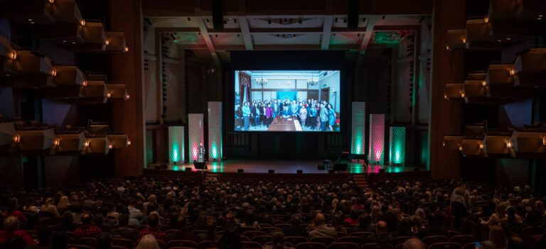A large audience sits in a theater, facing a stage with a giant screen displaying a group photo. The auditorium is dimly lit, with lights highlighting the stage and side walls.