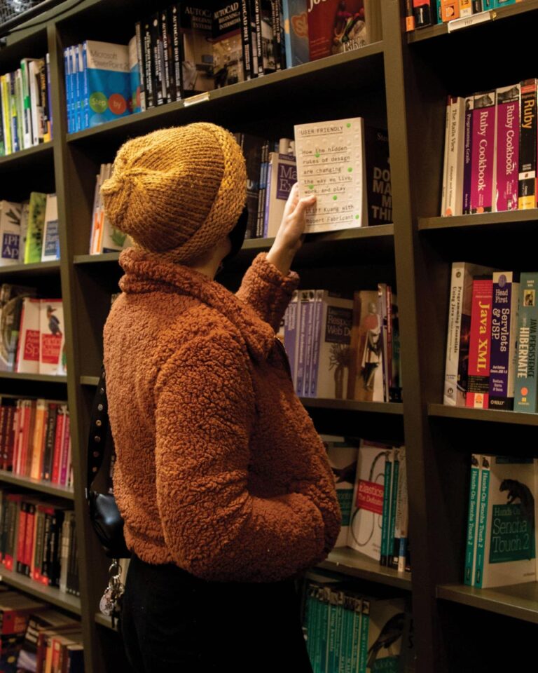 A person in a brown furry jacket and yellow beanie browses books in a bookstore. They are reaching for a book on a shelf filled with various titles.