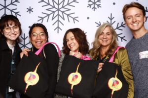 Five people smiling and holding black tote bags with a colorful "Respect" logo. They stand in front of a backdrop featuring large snowflake designs.