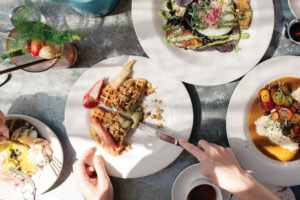 Five white plates with a variety of food on a grey table. Two hands hover above the plates. One hand holds a knife.