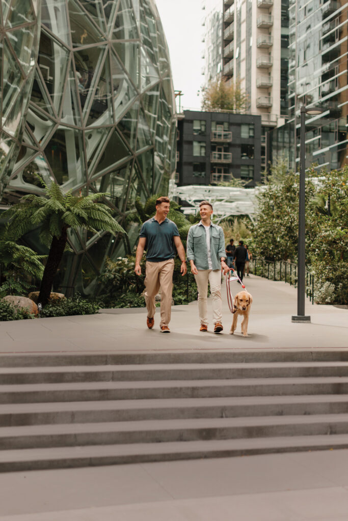 A photo of two people walking next to the Seattle Spheres. The person on the left has a short sleeve green polo and khaki pants on. The person on the right has a green long sleeve button up with a white t-shirt and khaki pants on. The person on the right holds the harness for a yellow colored guide dog.