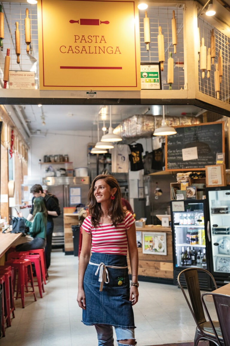 A woman stands smiling in a cozy pasta shop with wooden decor and rolling pins displayed above. She wears a striped shirt and denim apron. The shop, "Pasta Casalinga," features red stools, a menu board, and a refrigerator with drinks.