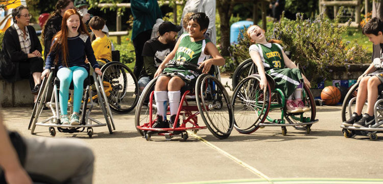 Four people in colorful clothing using wheelchairs on a paved court in the foreground with a crowd of people sitting on a bench in the background.
