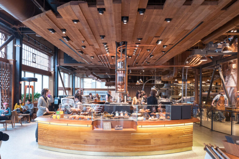 A bustling, modern café in Seattle features a large wooden counter displaying baked goods. Patrons order and work behind the counter while wooden panels and stylish lighting fixtures adorn the ceiling. Large windows flood the space with natural light, making it a must-visit option among things to do in Seattle.