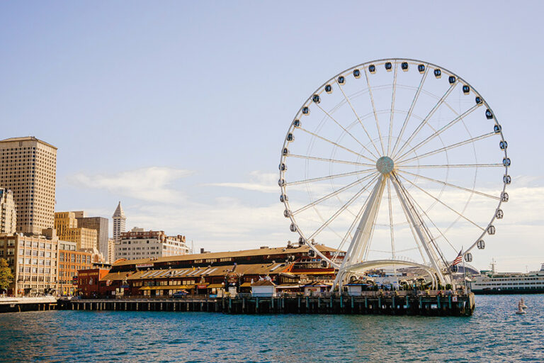 The Seattle Great Wheel on Pier 57 jets out into the blue waters of Elliott Bay with the Seattle skyline on the left.
