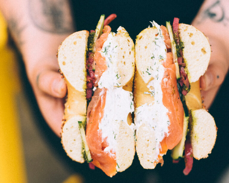 A close up of two hands holding a lox bagel sandwich that has been split down the center to display its contents.