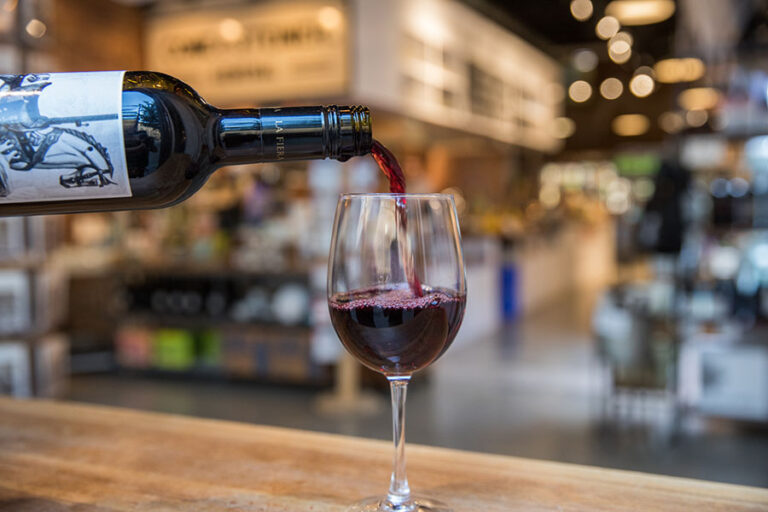 A bottle of red wine is being poured into a glass on a wooden counter. The background is a blurred interior of a cozy, warmly lit shop or cafe.