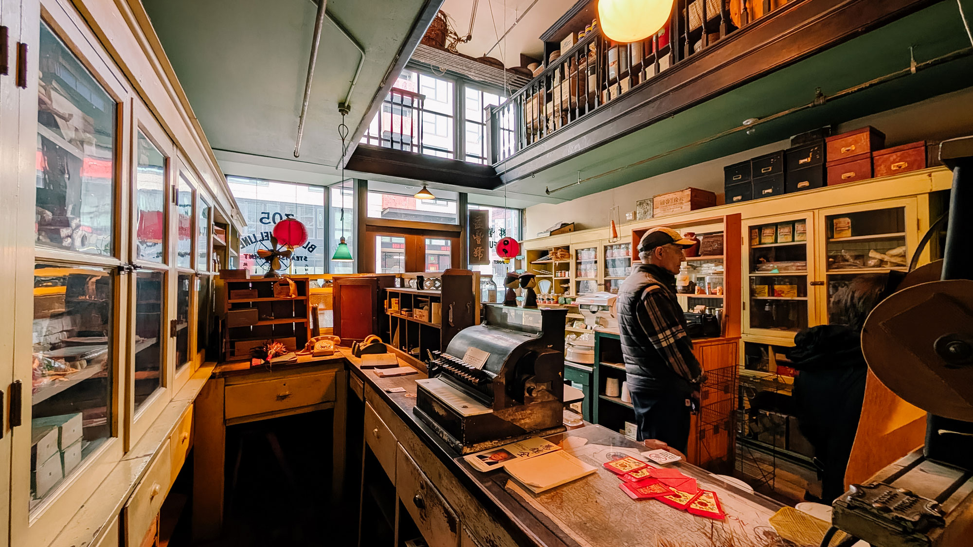 A vintage store interior with wooden counters, shelves filled with various items, and a cash register. A person in a cap stands near the back, and warm light filters through a large window. The space has a nostalgic, cluttered atmosphere.