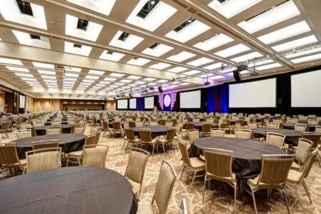 An empty conference hall with numerous round tables covered in dark tablecloths, surrounded by chairs. Large projection screens and stage lighting are visible at the front, suggesting a setup for a presentation or event.