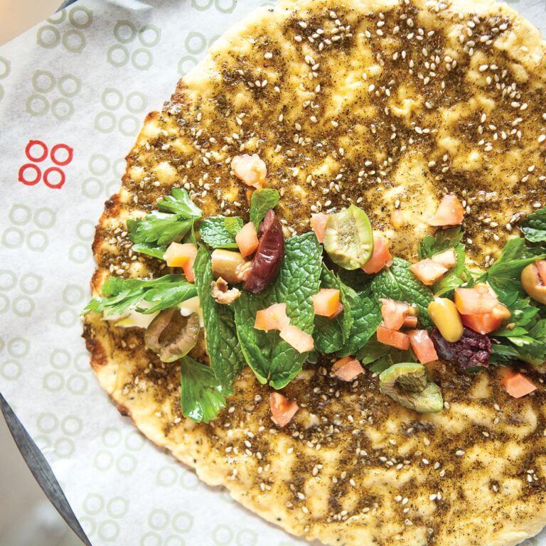 Flatbread topped with za'atar, fresh herbs, diced tomatoes, and assorted olives on a sheet of branded parchment paper.