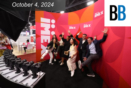 A group of people enthusiastically pose in front of a vibrant red and orange IMEX backdrop.