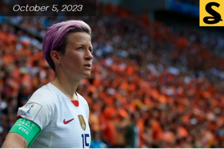 White middle aged women with short hair staring off. Wearing a soccer white shirt. A crowd of fans in the stands behind her.