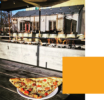 A plate with a slice of pizza on a wooden table outside a restaurant. The foreground shows a concrete ledge with decorative logs and the background features a row of barrels, patio lights, and a building with windows.