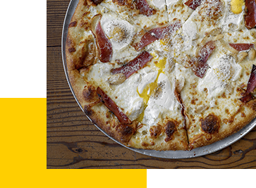 A pizza with a golden crust topped with white cheese, slices of prosciutto, and cracked eggs is served on a metal tray. It is placed on a wooden table with a yellow geometric design overlaying the corner of the image.